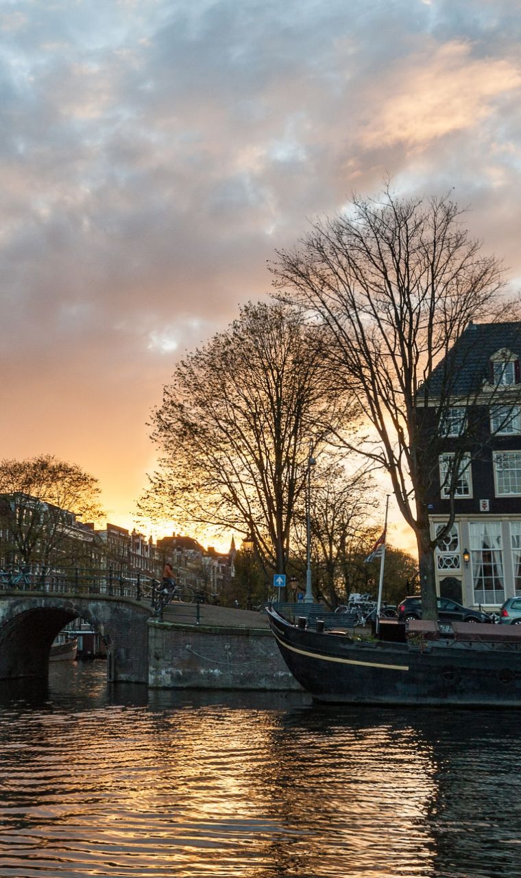 Zonsondergang boven een gracht in Amsterdam, met kale bomen, een brug en een woonboot
