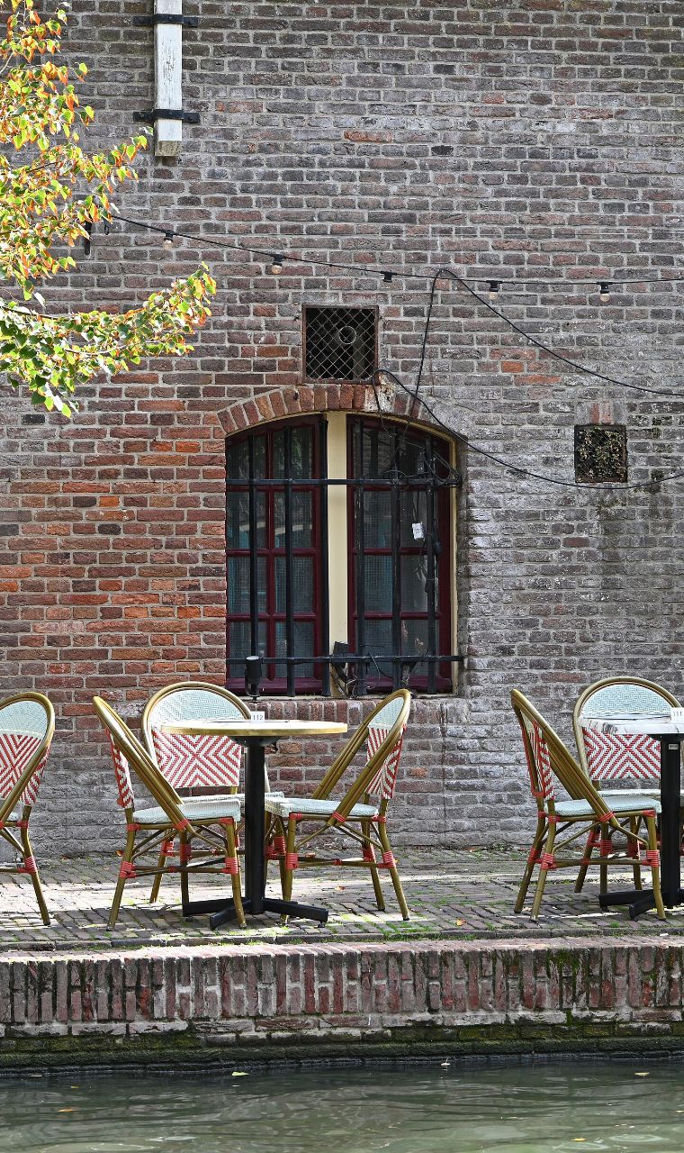 Een terras met rieten stoelen op een werf aan de Oudegracht in Utrecht, voor een bakstenen muur met een boogvenster
