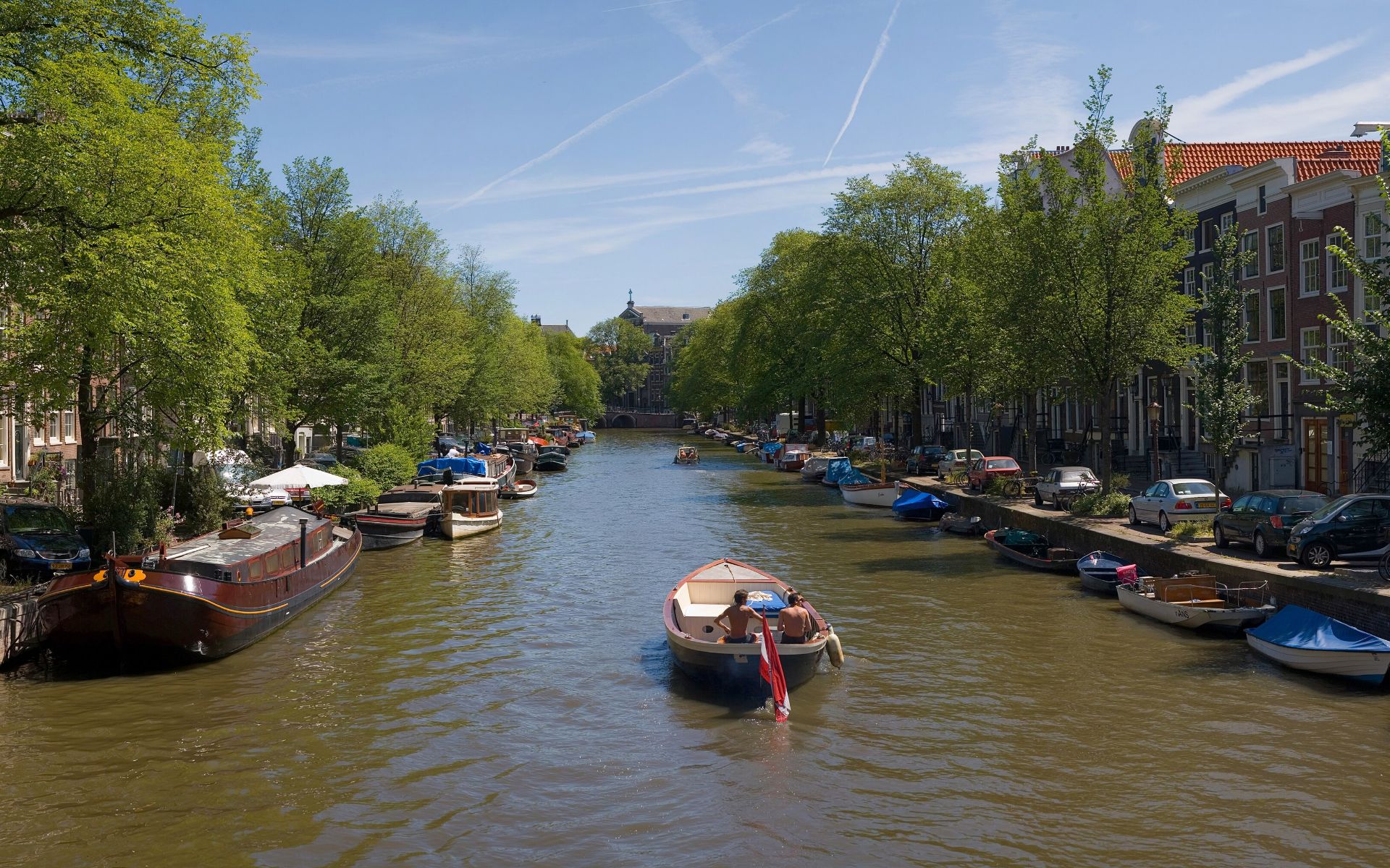 Een Amsterdamse gracht op een zomerdag: bomen langs de kade, woonboten, en een sloep met twee mensen, van achteren gezien